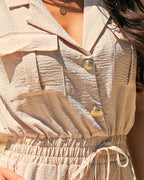 Light beige sleeveless notched collar jumpsuit with drawstring waist, flap pockets, and gold buttons.