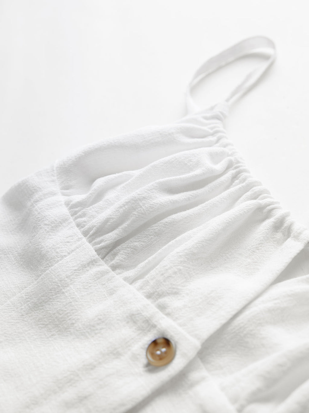 Soft white cotton mini dress with textured fabric, spaghetti straps, gathers, and brown button.