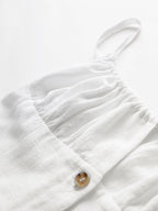 Soft white cotton mini dress with textured fabric, spaghetti straps, gathers, and brown button.