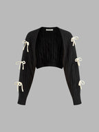 Black cropped cable knit cardigan with long sleeves and six cream ribbon bow accents.