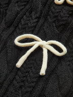 A cable knit cardigan with a cream-colored bow-knot accent and textured black knit pattern.
