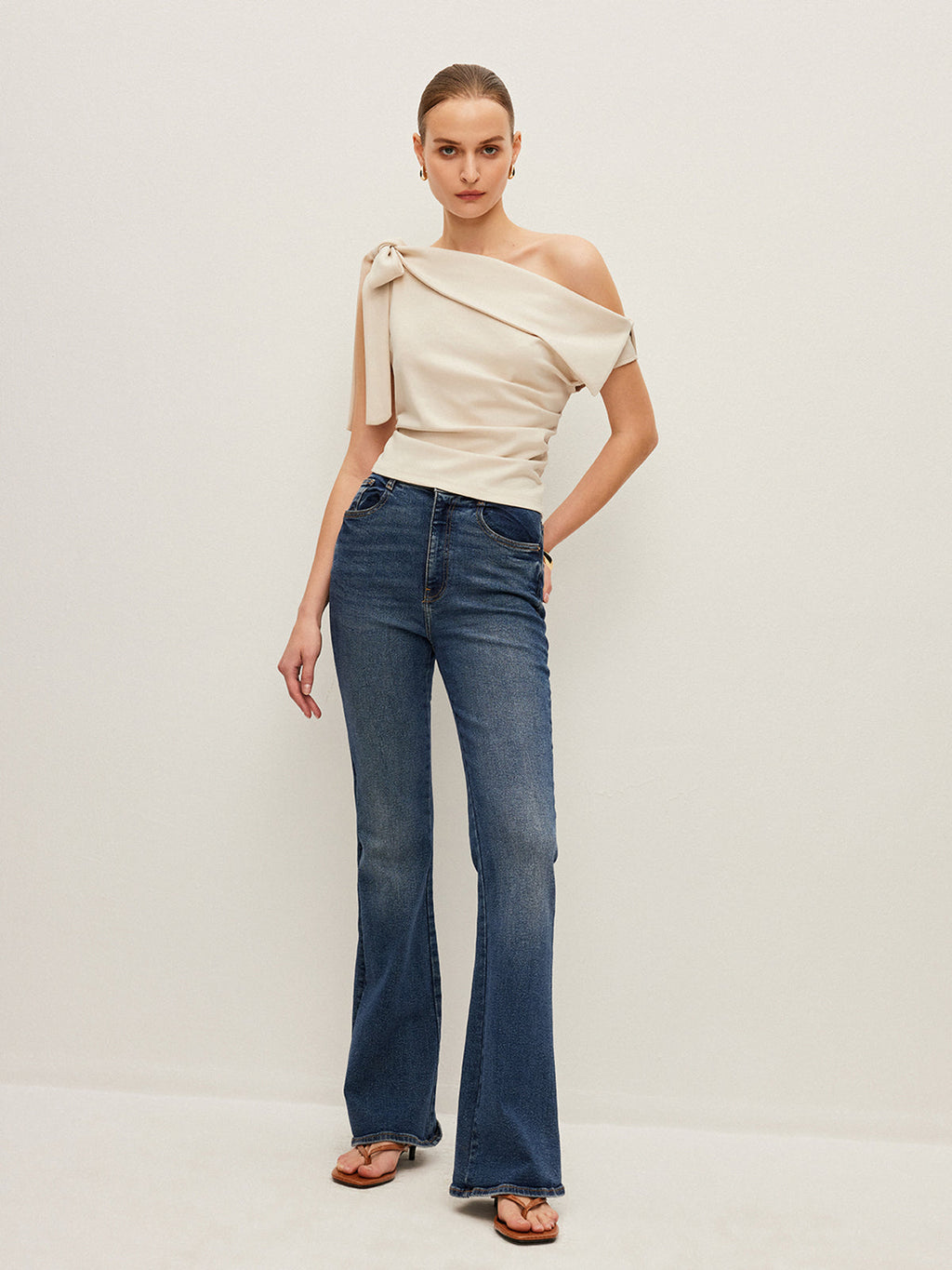 One-shoulder blouse with elegant bow detail, blue high-waisted flare jeans, brown sandals.
