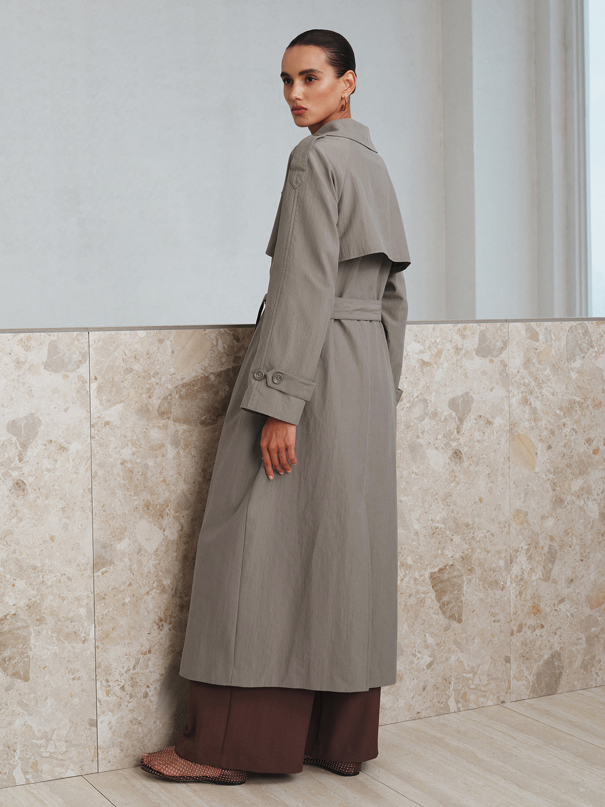 Grey belted cotton oversize trench coat with cape detail and buttoned cuffs, brown wide-leg trousers.