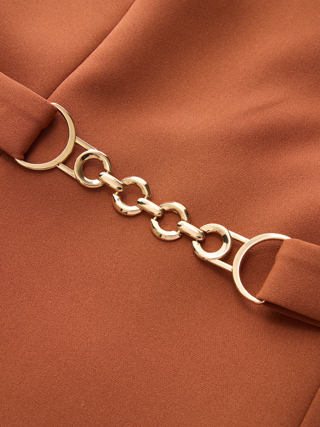 Rust-colored asymmetric neckline mini dress with belted waist, gold rings, and decorative chain detail.