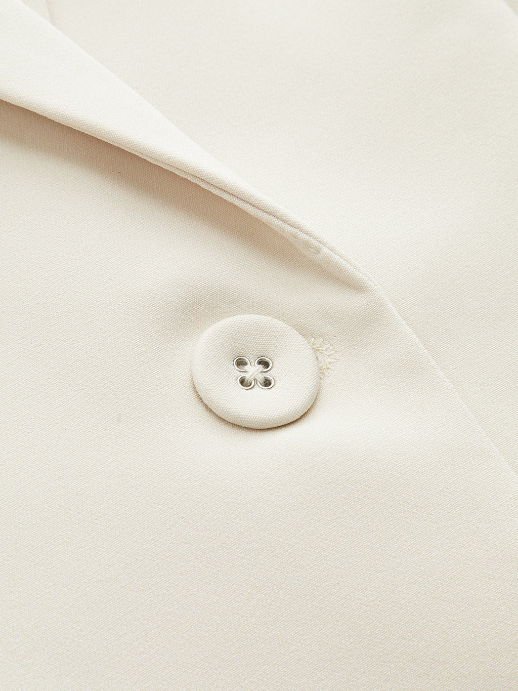 Cream blazer lapel with fabric-covered button and four silver eyelets, highlighting tailored texture.