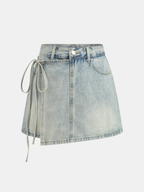 A denim mini skort with crossover front, side tie, asymmetric hem, faded wash, gold stitching.