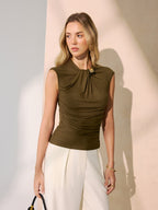 Olive green sleeveless blouse with twisted metal neckline, gold clasp, and ruched detailing; cream wide-leg trousers.