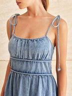 A denim midi dress with bow-tied adjustable straps, a fitted waist, and tiered gathered fabric.
